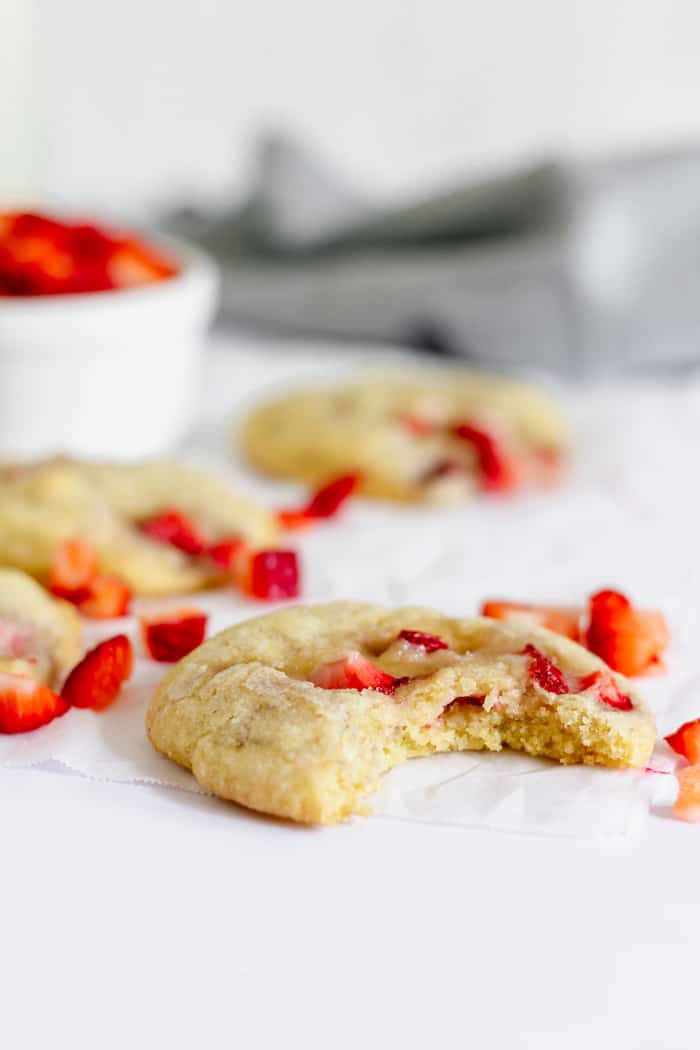 Image shows a strawberry chunk cookie with a bite taken out on a table, surrounded by small pieces of strawberry