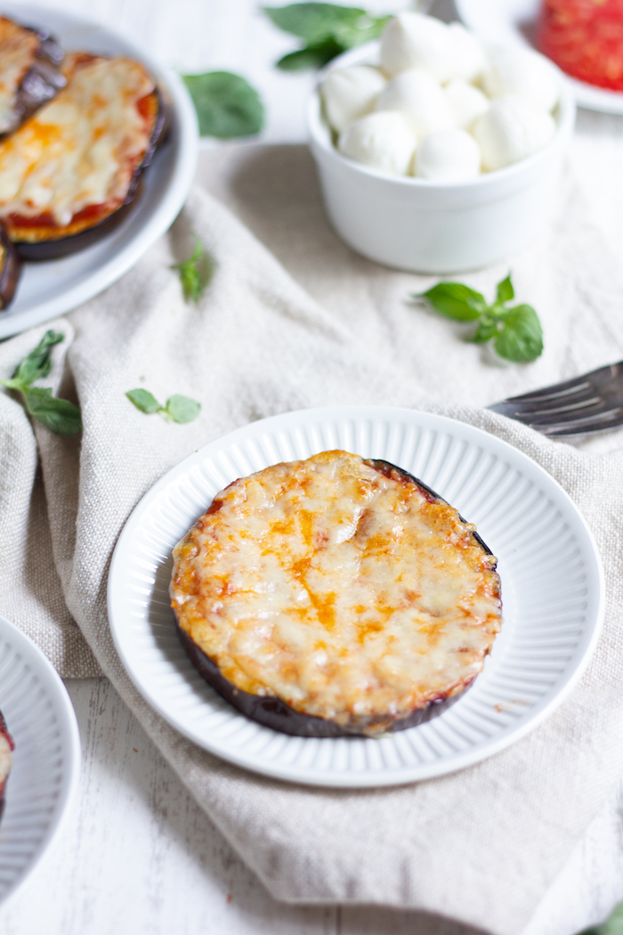 Photo depicts an eggplant pizza on a white plate in the foreground, with cheese and sauce in the background