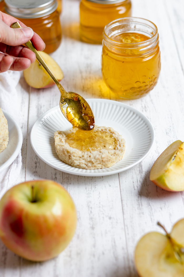 Image shows a small plate with half of a biscuit on it. A spoon is putting apple jelly on the biscuit. Nearby, apple slices and a jar of apple jelly sit on the table. 