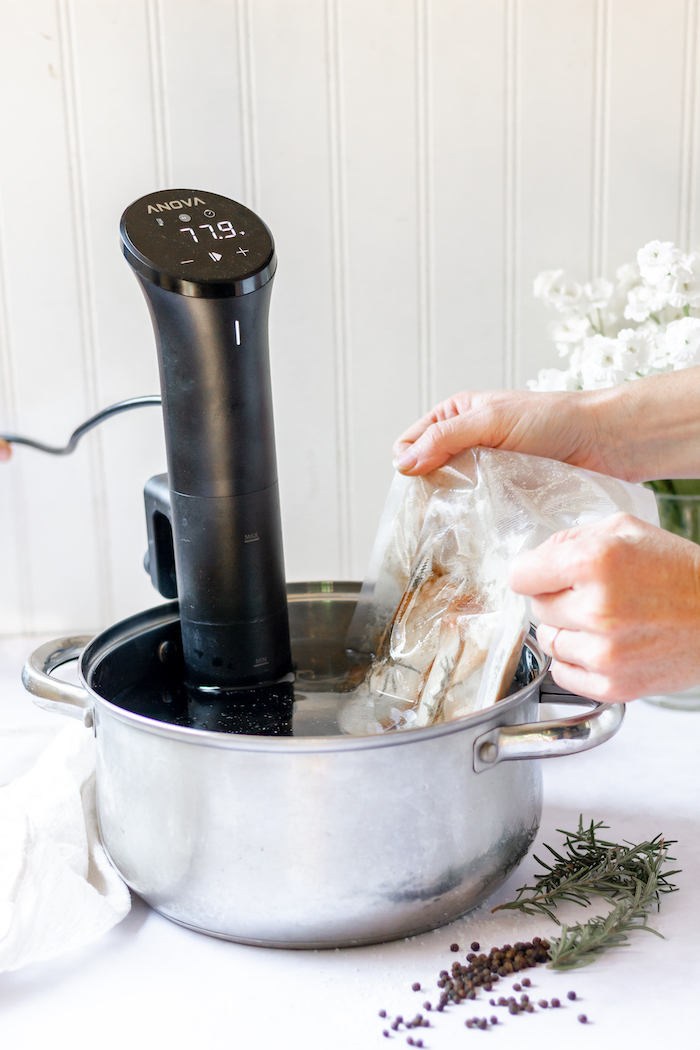 Image shows a pair of hands placing a pork loin into a large stock pot with a precision cooker immersed in the water. Next to the pot on the counter top sits some rosemary.