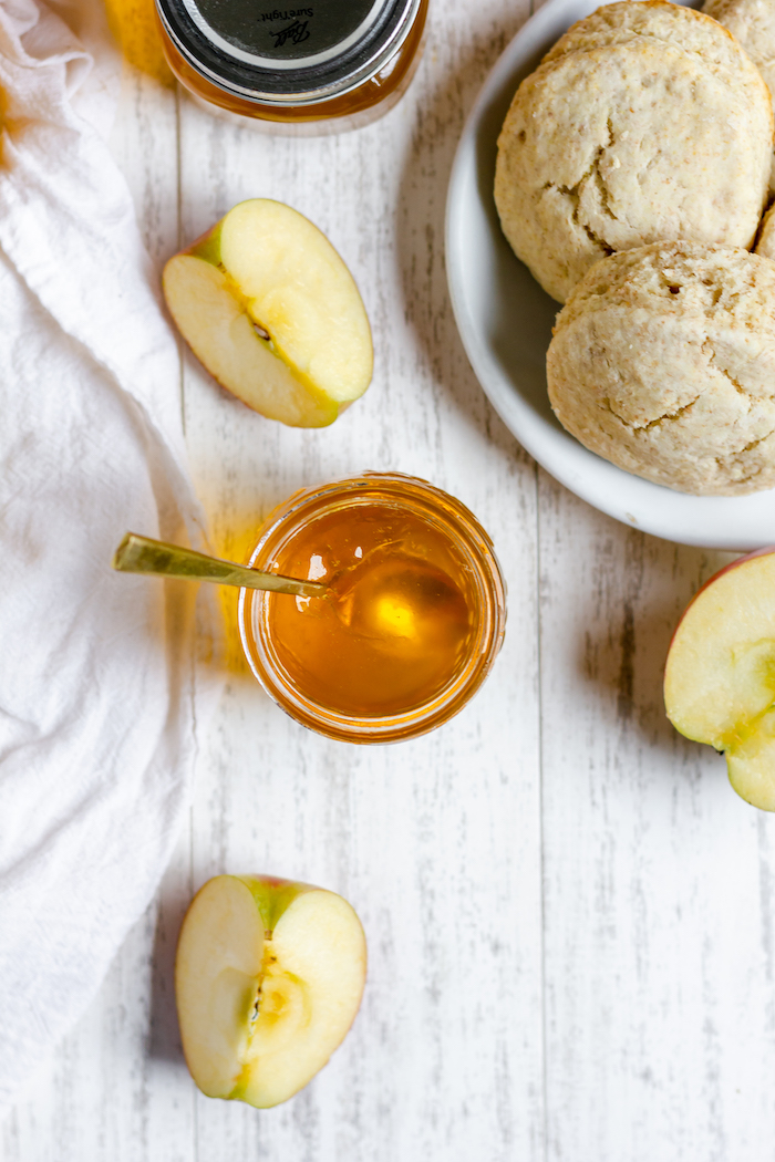 Image, taken from above, shows an open jar of apple jelly with a spoon in it. A few apple slices sit on the table nearby, along with a plate of biscuits. 