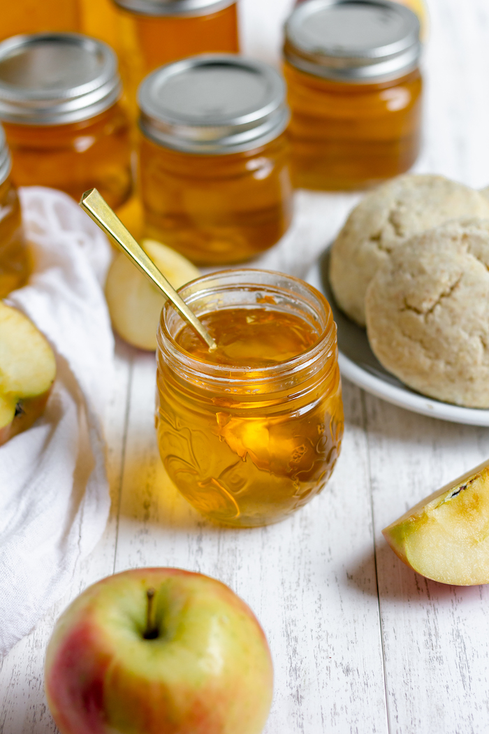 Image shows several jars of apple jelly lined up on a table behind a plate of rolls. A jar of open apple jelly with a spoon in it sits at the front of the photo. 