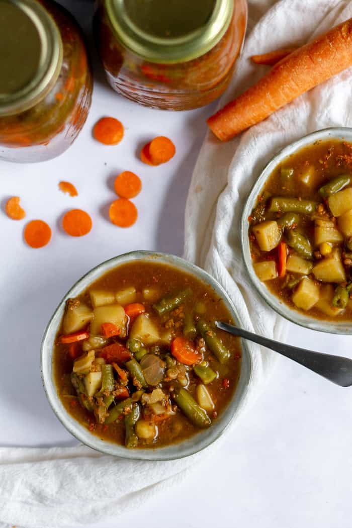 Image, taken from above, shows two bowls of hearty, vegetable soup, one with a spoon in it. Nearby sit two jars of more soup, and a carrot. 