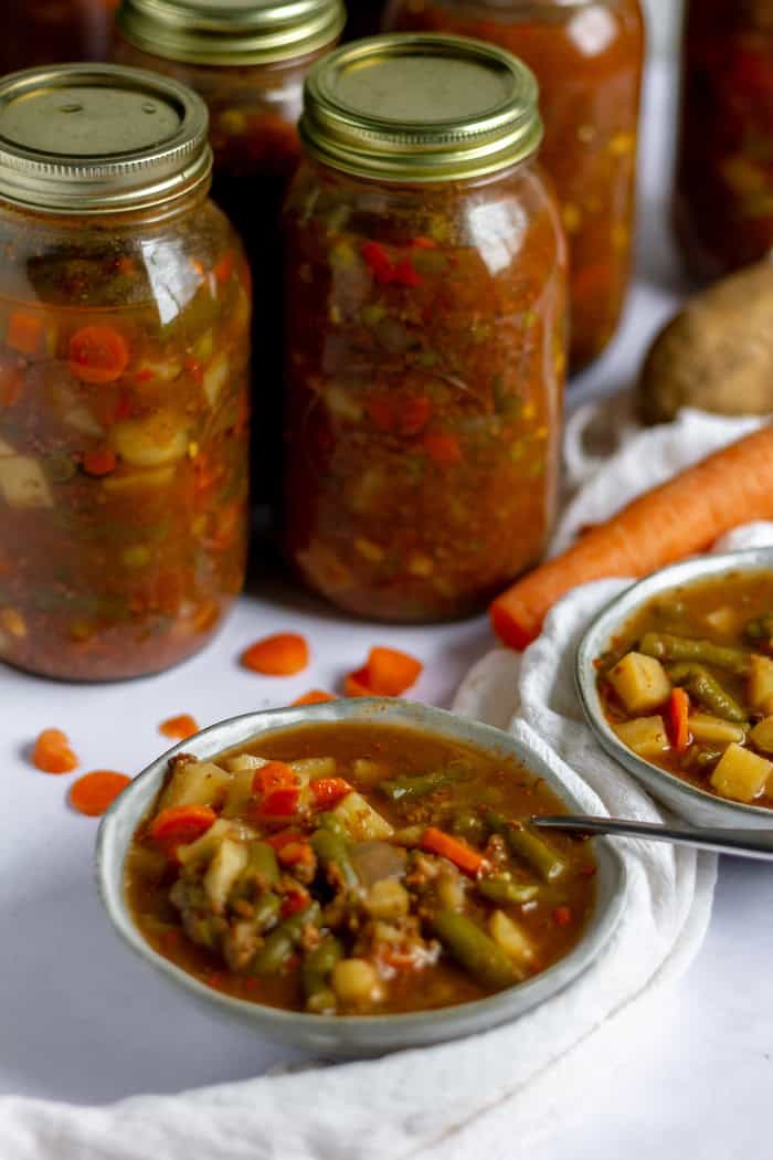 Image shows several jars of vegetable soup in the background, with two bowls of soup on the counter in front. A spoon rests in one of the bowls. 
