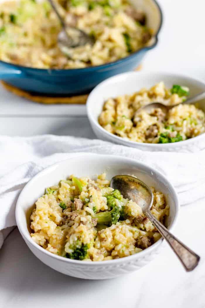 Photo shows a bowl of cheesy broccoli rice skillet in the foreground, with another bowl and a blue skillet of the rest of the meal in the background. 