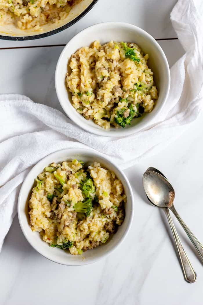 Image, taken from above, shows two white bowls on a white tablecloth, each with a serving of cheesy broccoli rice skillet meal. Next to the bowls sits two silver spoons. 
