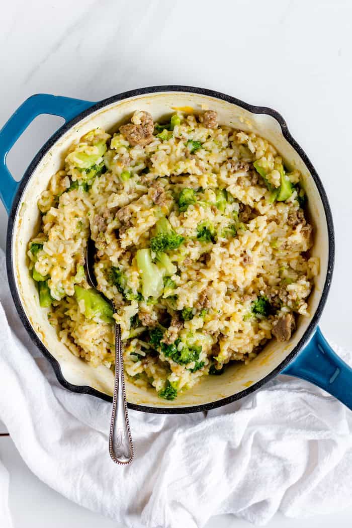 Image, taken from above, shows a large blue skillet full of cheesy broccoli rice casserole with a spoon in it to serve with. 