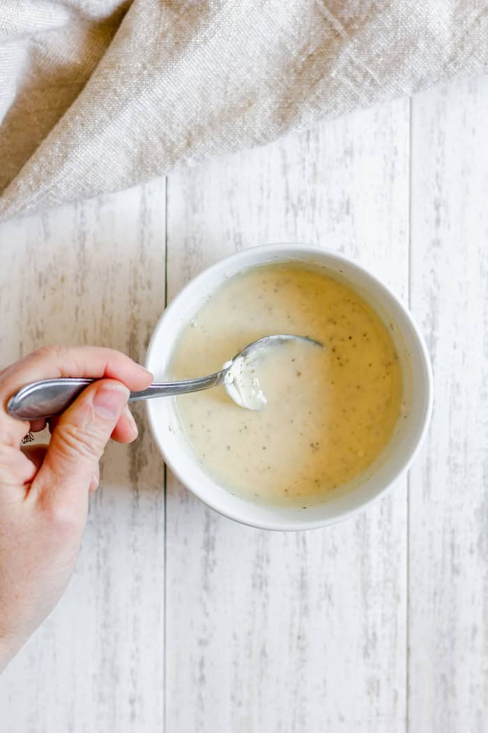Image, taken from above, shows a hand holding a spoon in a while bowl of dressing, having mixed all the ingredients to make dressing for broccoli bacon salad.