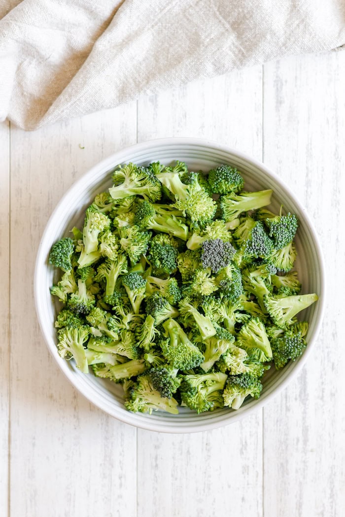 Image, taken from above, shows a large white bowl of cut broccoli sitting on a table.