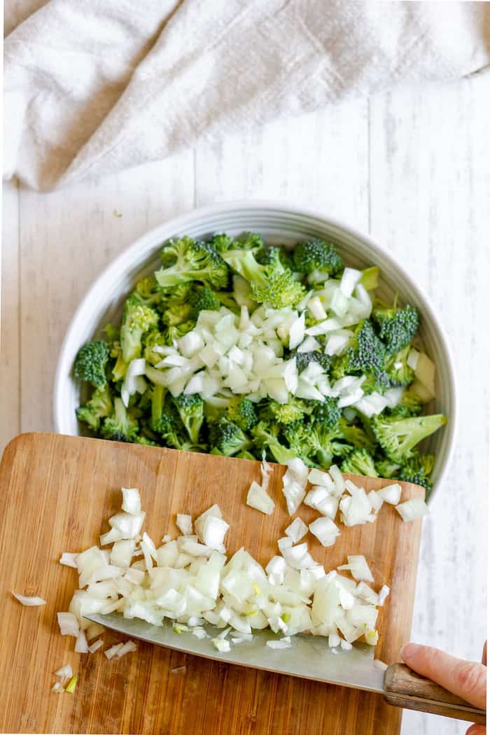 Image, taken from above, shows a large white bowl of cut broccoli with a knife adding onions from a cutting board.