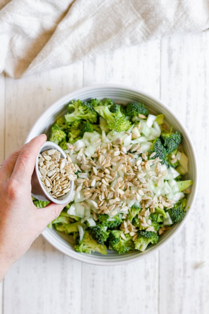 Image, taken from above, shows a large white bowl of broccoli and onions, with a hand pouring a small bowl of sunflower seeds into the large bowl.