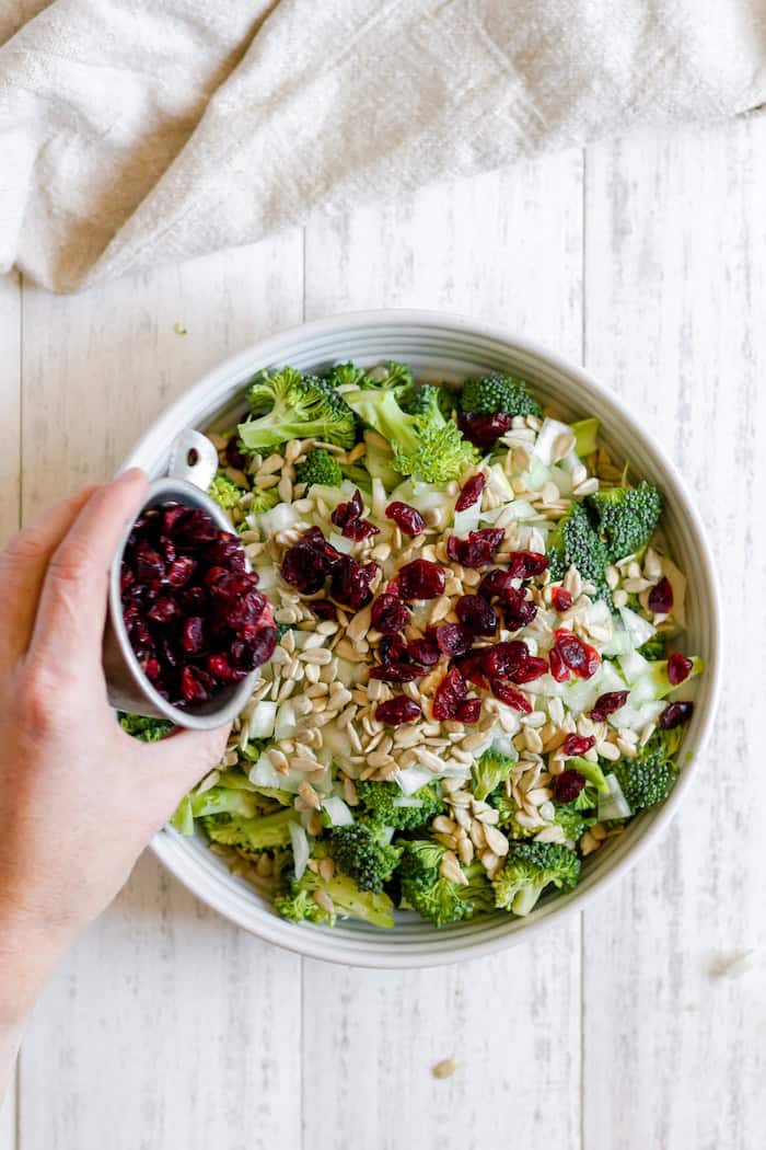 Image, taken from above, shows a hand pouring a small bowl of craisins into a large bowl of broccoli bacon salad.