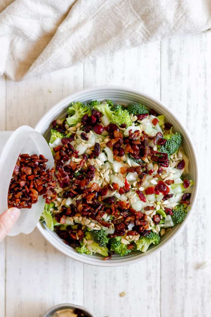 Image, taken from above, shows a hand pouring a small dish of bacon into a large white bowl of broccoli bacon salad ingredients.