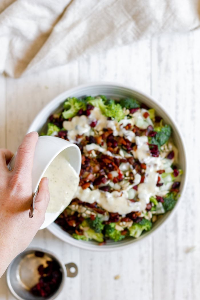 Image, taken from above, shows a hand pouring a dish of dressing into the large bowl of broccoli bacon salad.