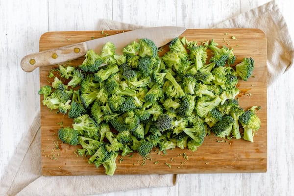 Image, taken from above, shows chopped broccoli on a cutting board with a knife, ready to make broccoli bacon salad.