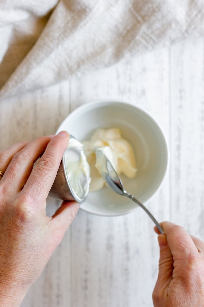 Image, taken from above, shows hands spooning mayo into a small white bowl, which sits on a white towel.