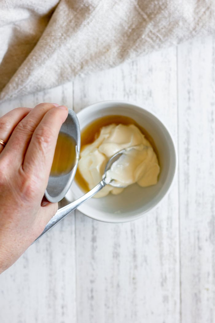 Image, taken from above, shows hands pouring apple cider vinegar into a small white bowl of mayo, which sits on a white towel.