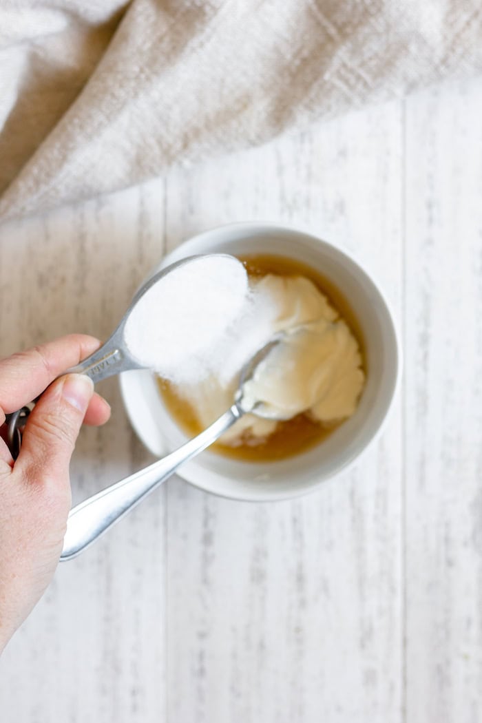Image, taken from above, shows hands pouring sugar into a small white bowl of mayo and apple cider vinegar, which sits on a white towel.