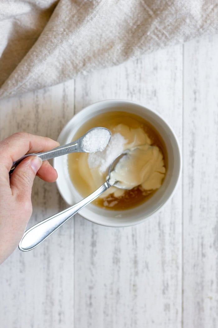 Image, taken from above, shows hands pouring salt into a small white bowl of mayo, apple cider vinegar and sugar, which sits on a white towel.