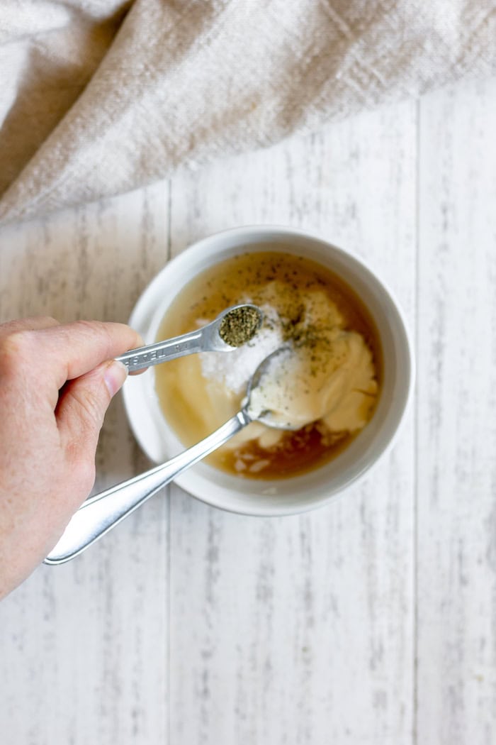 Image, taken from above, shows a hand pouring ground black pepper into a small white bowl of mayo, vinegar, sugar and salt, while sitting on a white table.