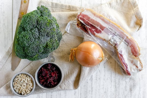 Image, taken from above, shows the ingredients for broccoli bacon salad laid out on a towel. Broccoli, bacon, onion, craisins and sunflower seeds.