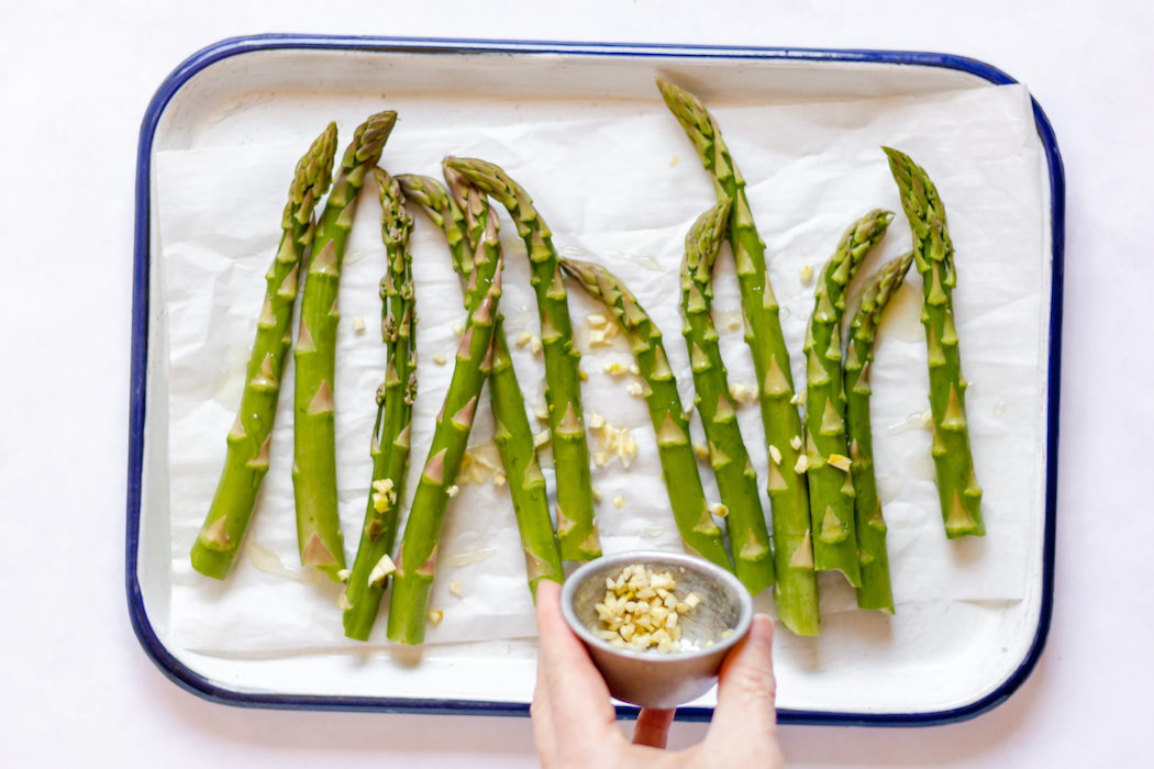 Image, taken from above, shows a tray of asparagus and a hand sprinkling diced garlic on the stalks.