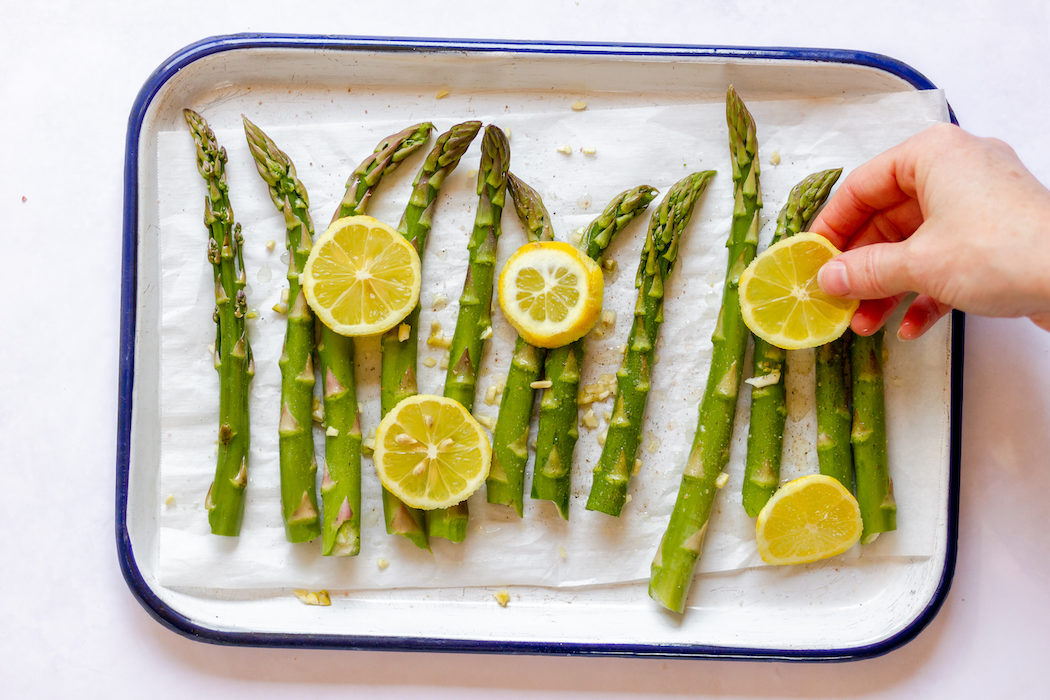 Image, taken from above, shows a hand placing lemon slices on top of asparagus stalks and garlic.