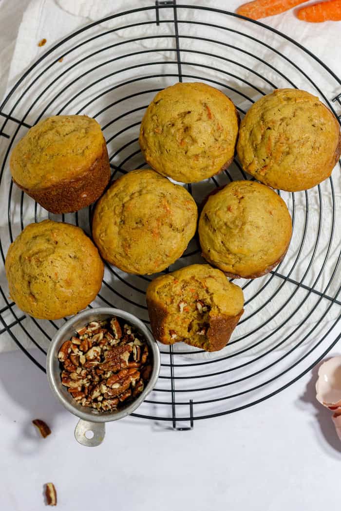 Photo, taken from above, shows seven gluten free carrot muffins on a wire trivet. One muffin has a bite taken out of it, and a small bowl of walnuts sits nearby.