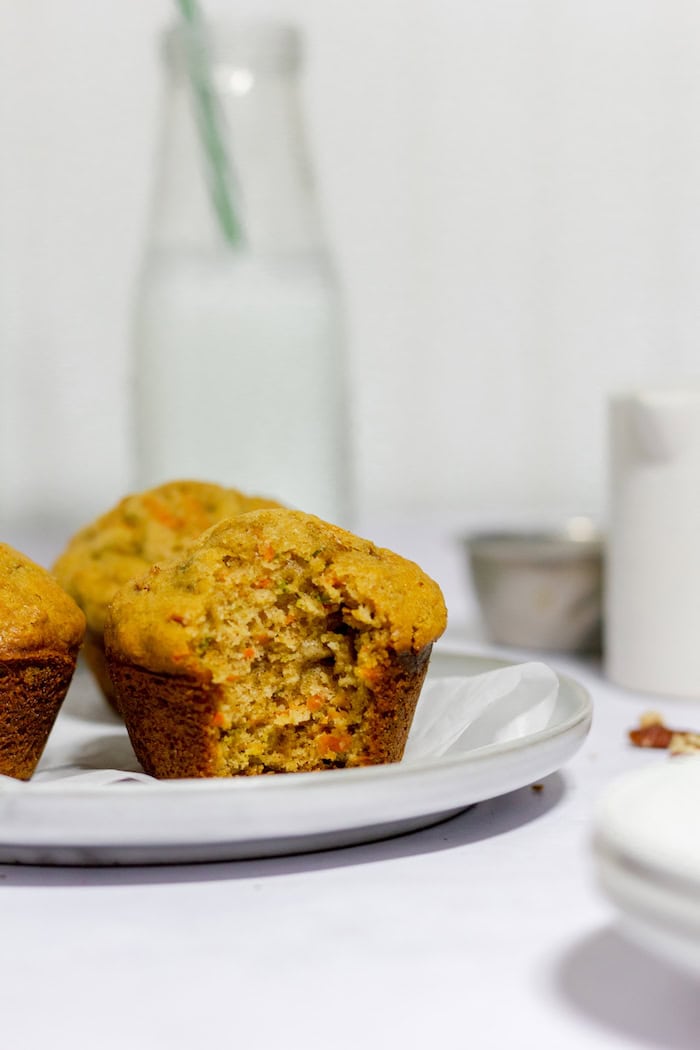 Photo shows three gluten free carrot muffins sitting on a white plate in a white kitchen. Front muffin has a bite taken out of it.