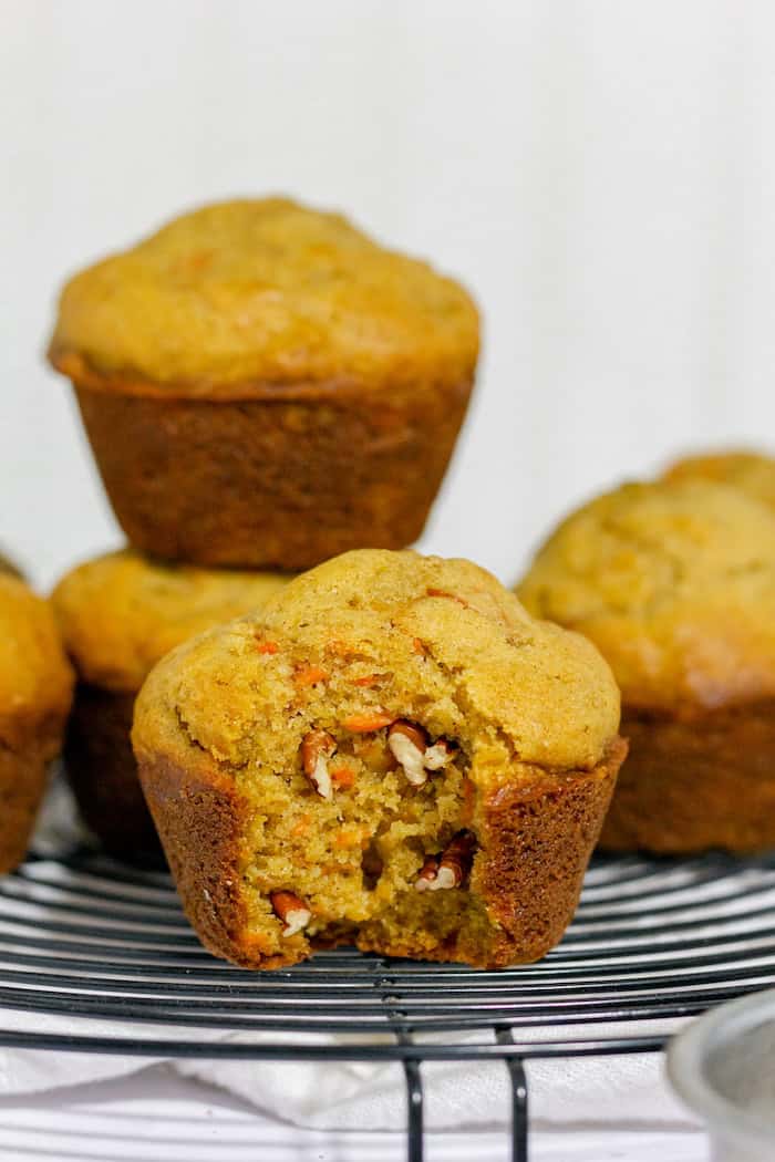 Image shows a close up of gluten free carrot muffins on a cooling rack, with one stacked on another in the background. Front muffin has a small bite taken out of it, showing pecans inside.