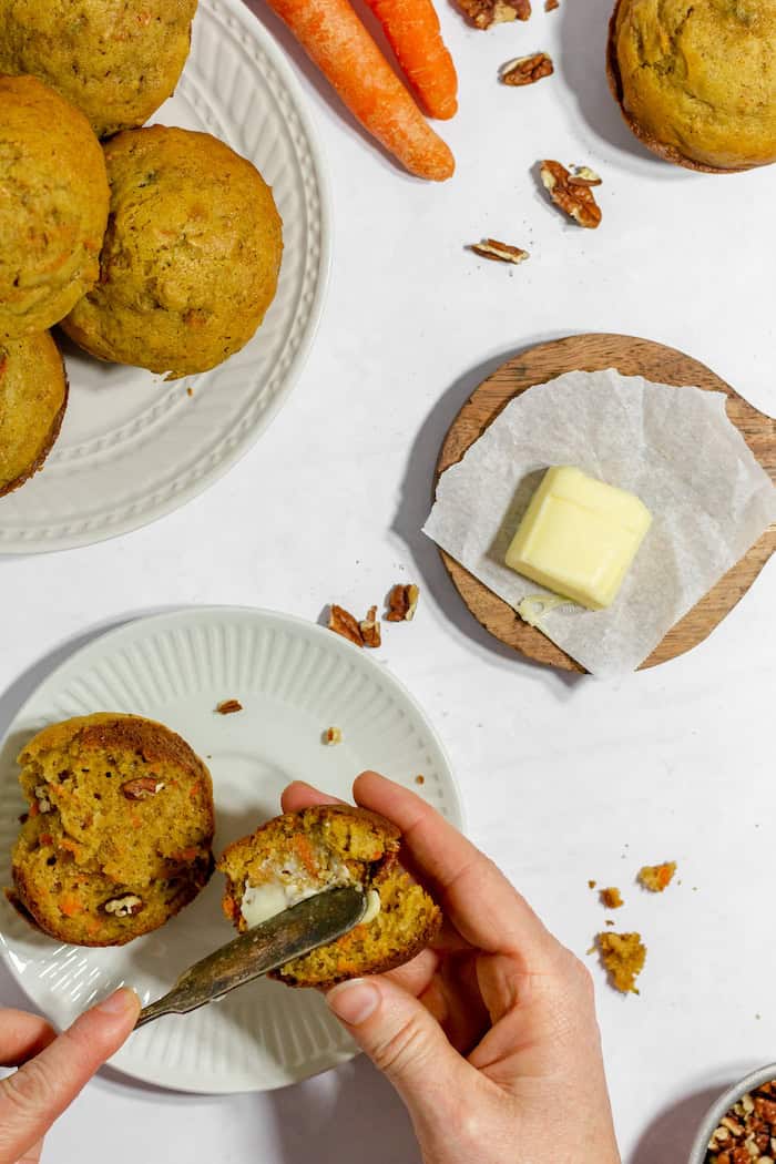 Image, taken from above shows a woman's hands spreading butter onto a gluten free carrot muffin. Nearby sits a wooden plate with more butter, and a white plate filled with more muffins.