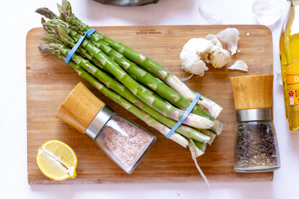 Images shows a bundle of asparagus on a cutting board with salt, pepper and lemon nearby.