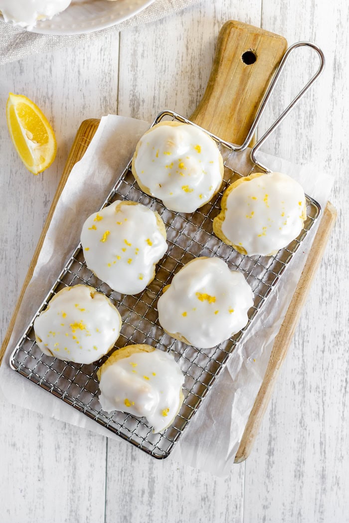 Image shows a wooden cutting board on a table, with a cooling rack holding a half dozen Italian Lemon Cookies on it. Next to the board sits a wedge of lemon. 