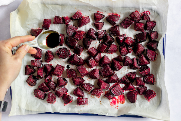 Image, taken from above, shows a cookie sheet with beets spread out for roasting. A hand drizzles balsamic over them.