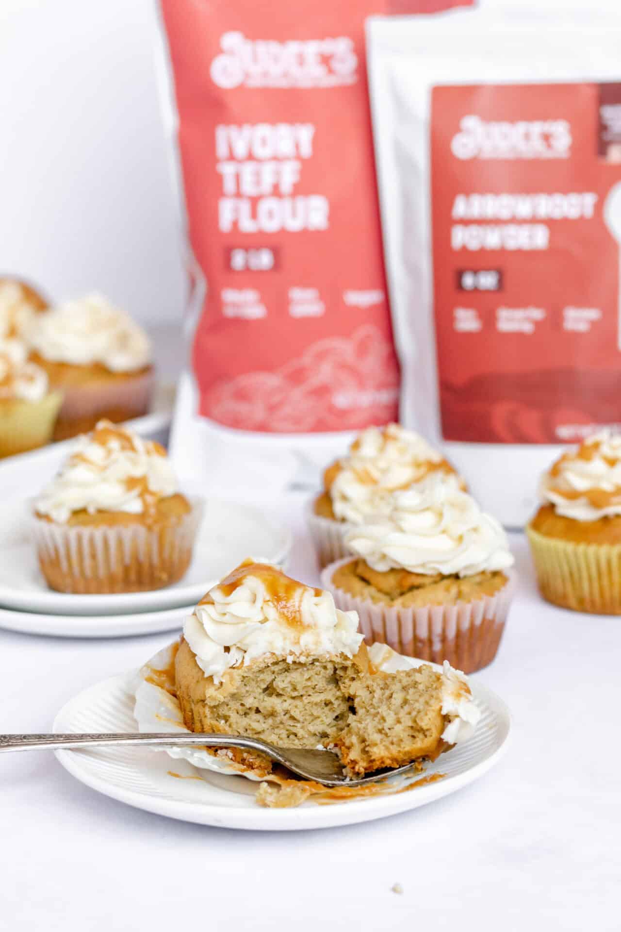 Image shows several gluten free banana cupcakes on plates and a countertop. The front cupcake has a bite cut into it with a fork. In the background are several Judee's gluten free products to bake with. 