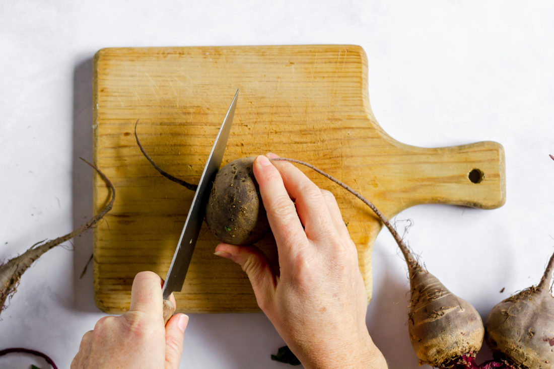 Image, taken from above, shows a hand slicing into a beet on a wooden cutting board with a large knife. 