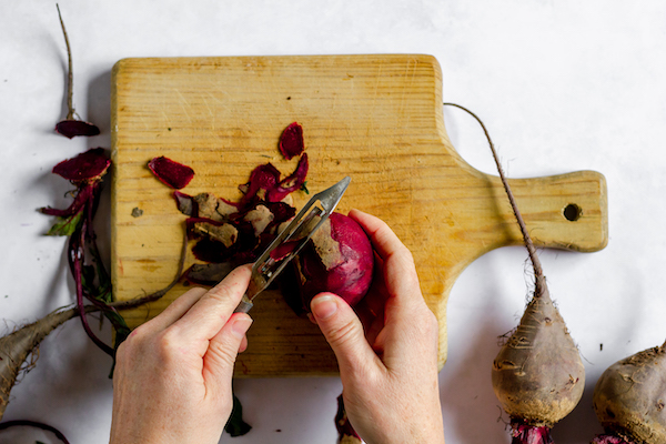 Image, taken from above, shows hands peeling the outer layer off a beet over a cutting board.