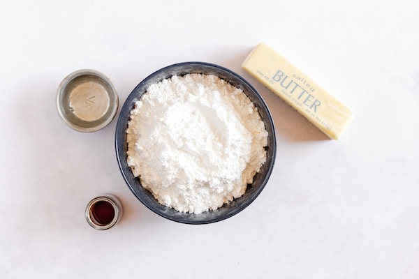 Image, taken from above, shows a small mixing bowl with powdered sugar in it, nearby sit butter, vanilla and water.