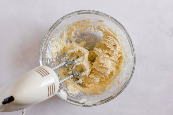 Image shows a glass mixing bowl with the butter and powdered sugar mixed together. A mixer rests on the side of the bowl. 