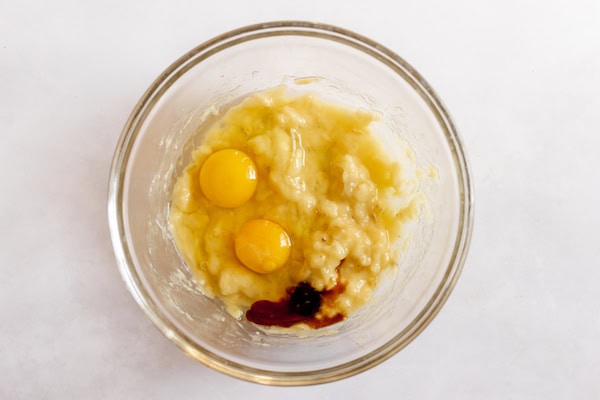 Image, taken from above, shows a glass bowl with mashed banana batter, plus eggs and vanilla. 