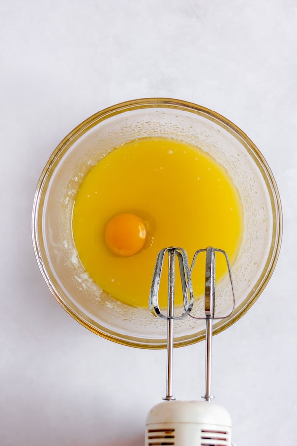 Image, photographed from above, shows an egg yolk floating in a glass bowl full of melted butter and sugar, with beaters resting over it.