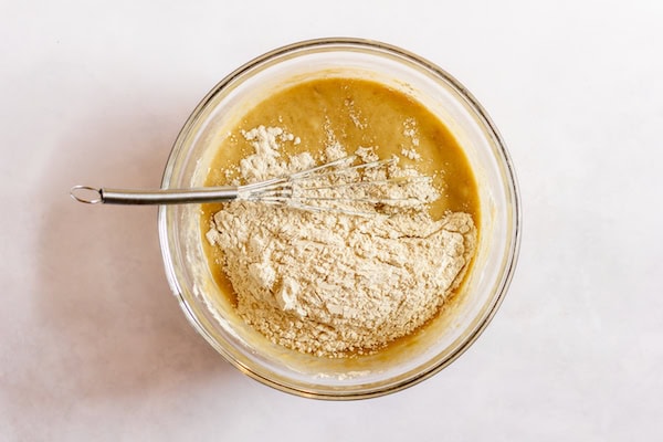 Image, taken from above, shows a glass bowl with batter for banana cupcakes, with flours being added in and a whisk resting in the bowl. 