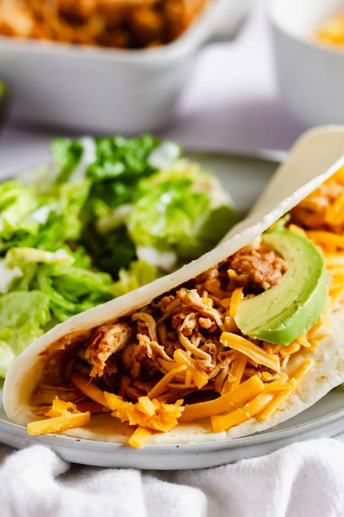 Image shows a close up of a chicken taco with shredded meat and cheese on a plate next to a bed of lettuce.