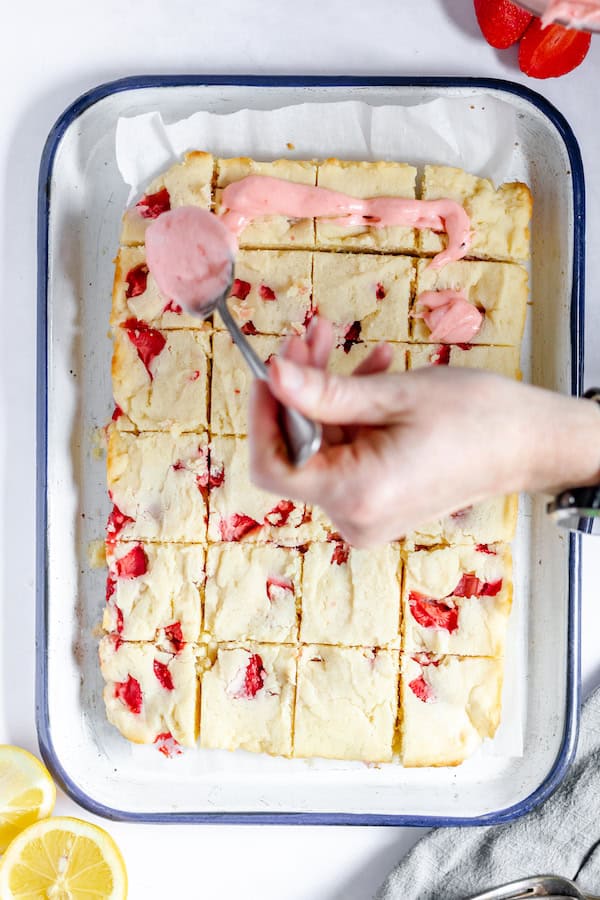 Photo, taken from above, shows a hand drizzling strawberry icing over the strawberry lemon blondies in a white pan.
