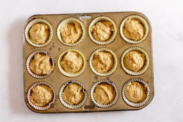 Image, taken from above, shows a muffin tin with 12 lined cups with batter for banana cupcakes in each cup. 