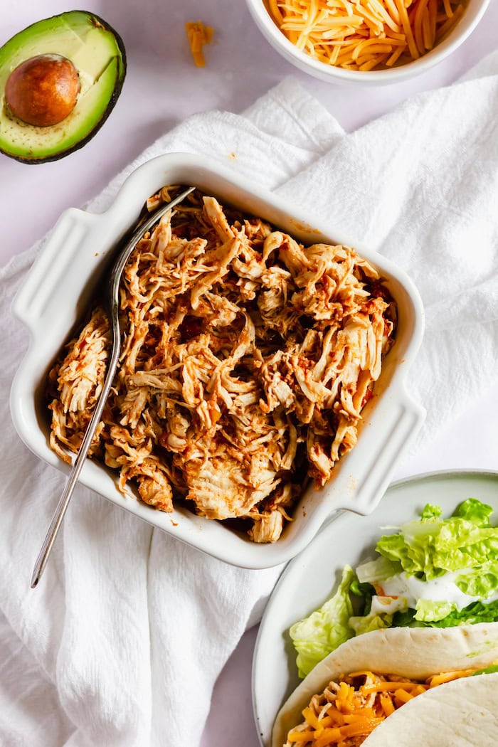 Image shows a table, photographed from above, with a square dish of shredded chicken with a spoon in it. Next to the dish on the table is a bowl of shredded cheese, half an avocado, and plate with a chicken taco on it. 
