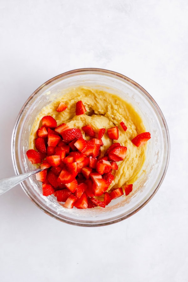 Images, taken from above, shows sliced strawberries in a glass bowl with a spoon and batter, ready to be folded into the batter.