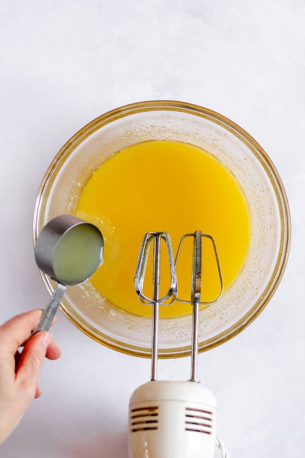 Image, taken from above, shows a hand pouring lemon juice into a bowl of melted butter, egg and sugar, with beaters resting on the side of the bowl.