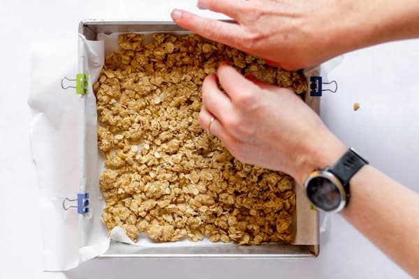 Image, taken from above, shows a hand spreading the granola mixture throughout a square pan. 