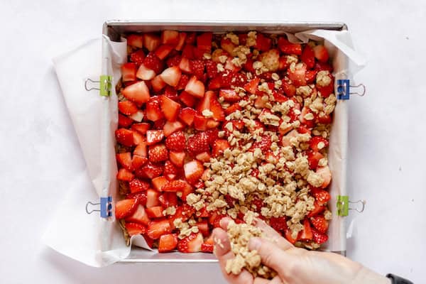 Image, taken from above, shows a hand sprinkling oatmeal granola in a pan over strawberries. 